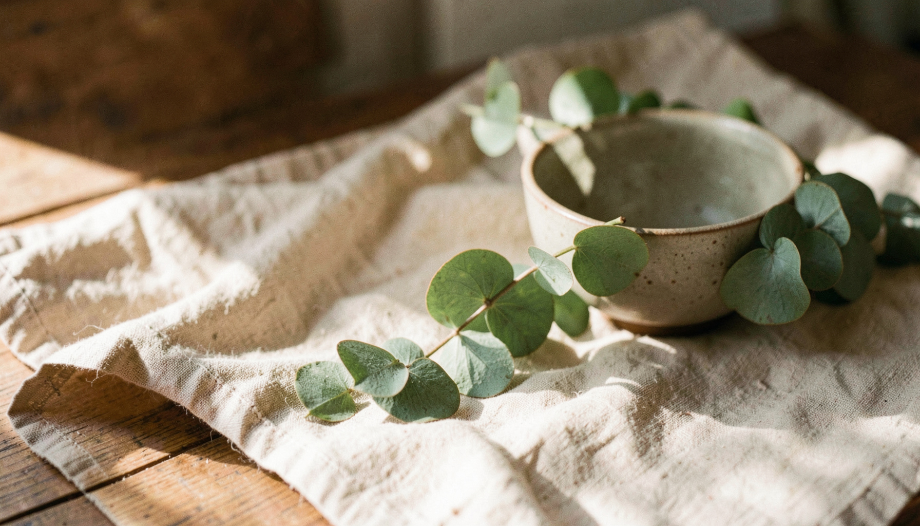 Soft natural light falling across a wooden surface with a linen cloth, a ceramic bowl, and a sprig of fresh eucalyptus, in warm earthy tones